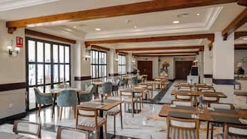 Un vaste espace dédié à la salle à manger, avec des tables et des chaises en bois, de grandes fenêtres et un plafond haut avec des poutres apparentes.