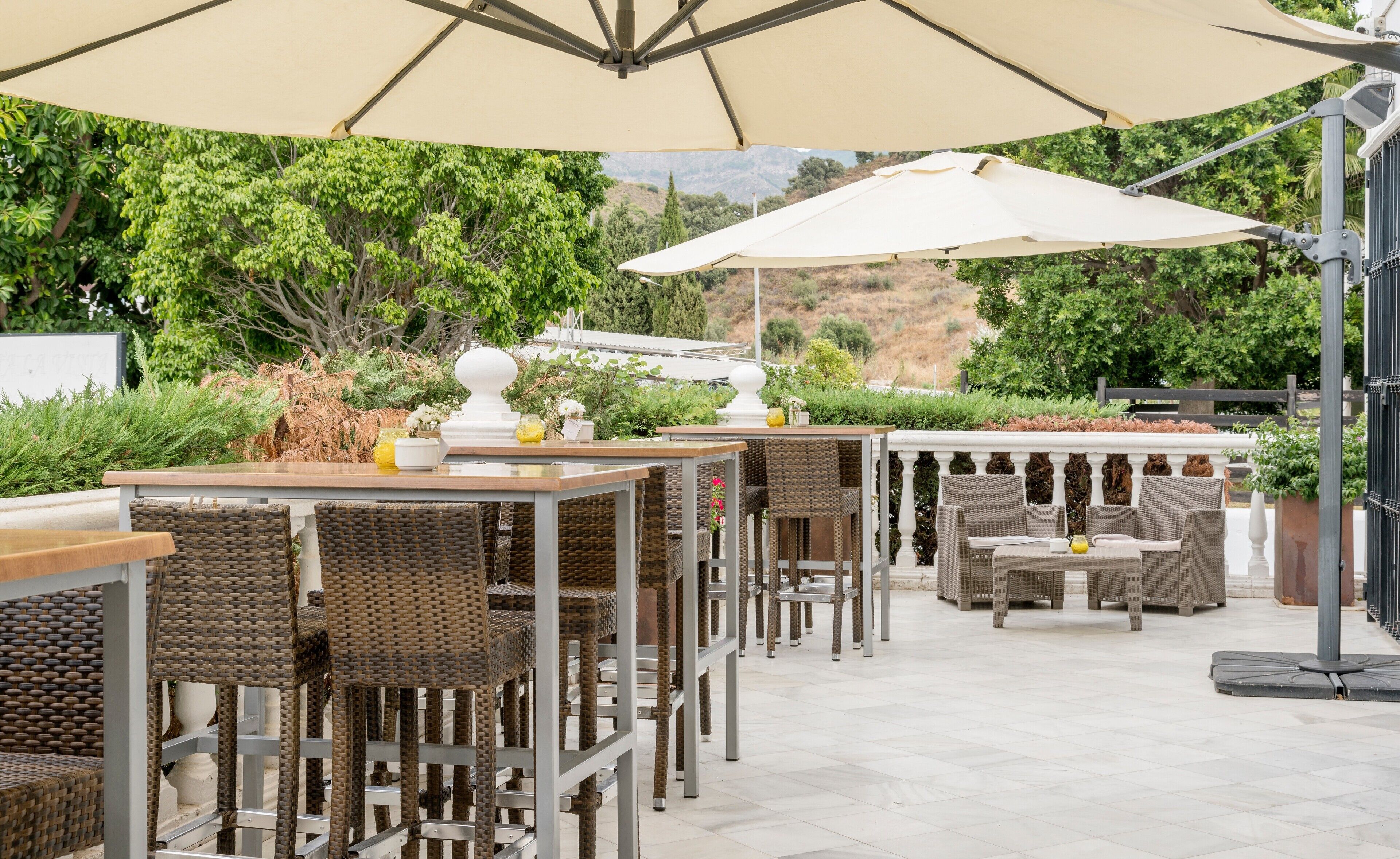 An outdoor patio with wicker furniture, beige umbrellas, and a view of greenery and hills.
