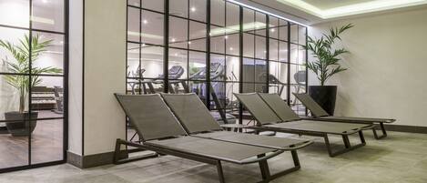 A modern indoor area with three lounge chairs, a large potted plant, and exercise equipment visible through glass walls.