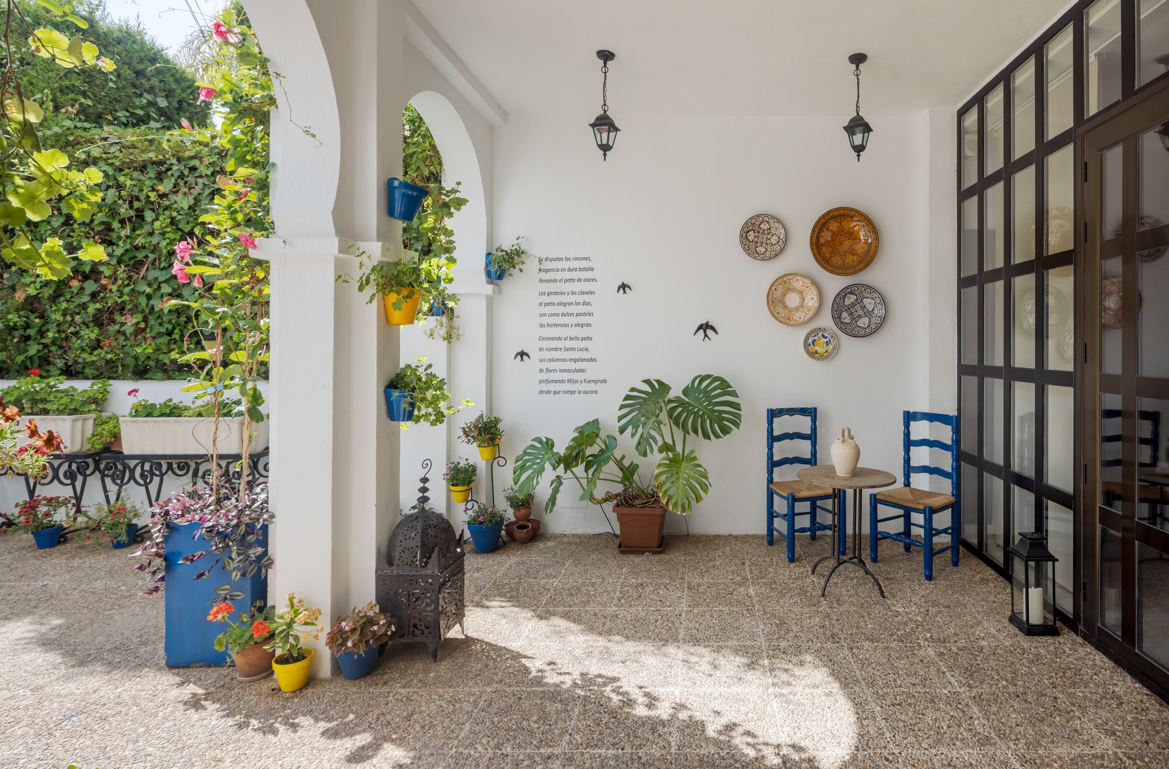 Ein überdachter Patio-Bereich mit Blumentöpfen, einem Tisch mit Stühlen und dekorativen Tellern an der Wand.