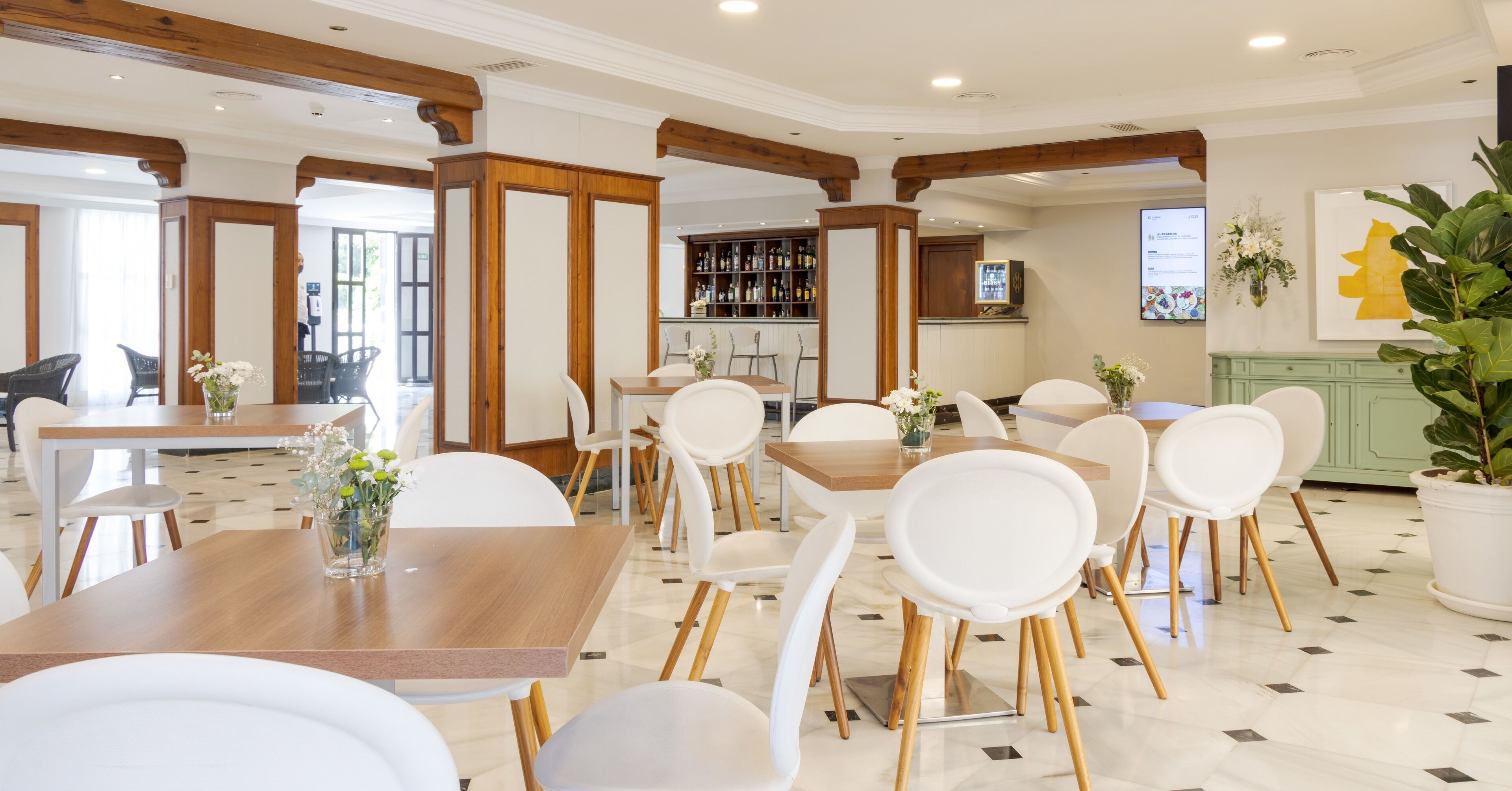 Un'ampia sala da pranzo con sedie bianche e tavoli di legno, una zona bar con diverse bottiglie e una grande pianta in vaso.