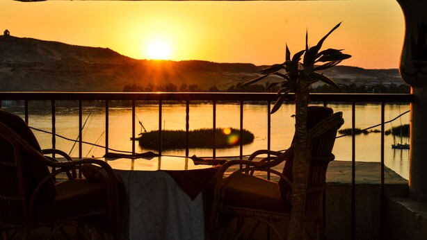 A balcony with wicker chairs and a table, overlooking a body of water with a sunset in the background. - Marhaba Hotel Aswan (Aswan)