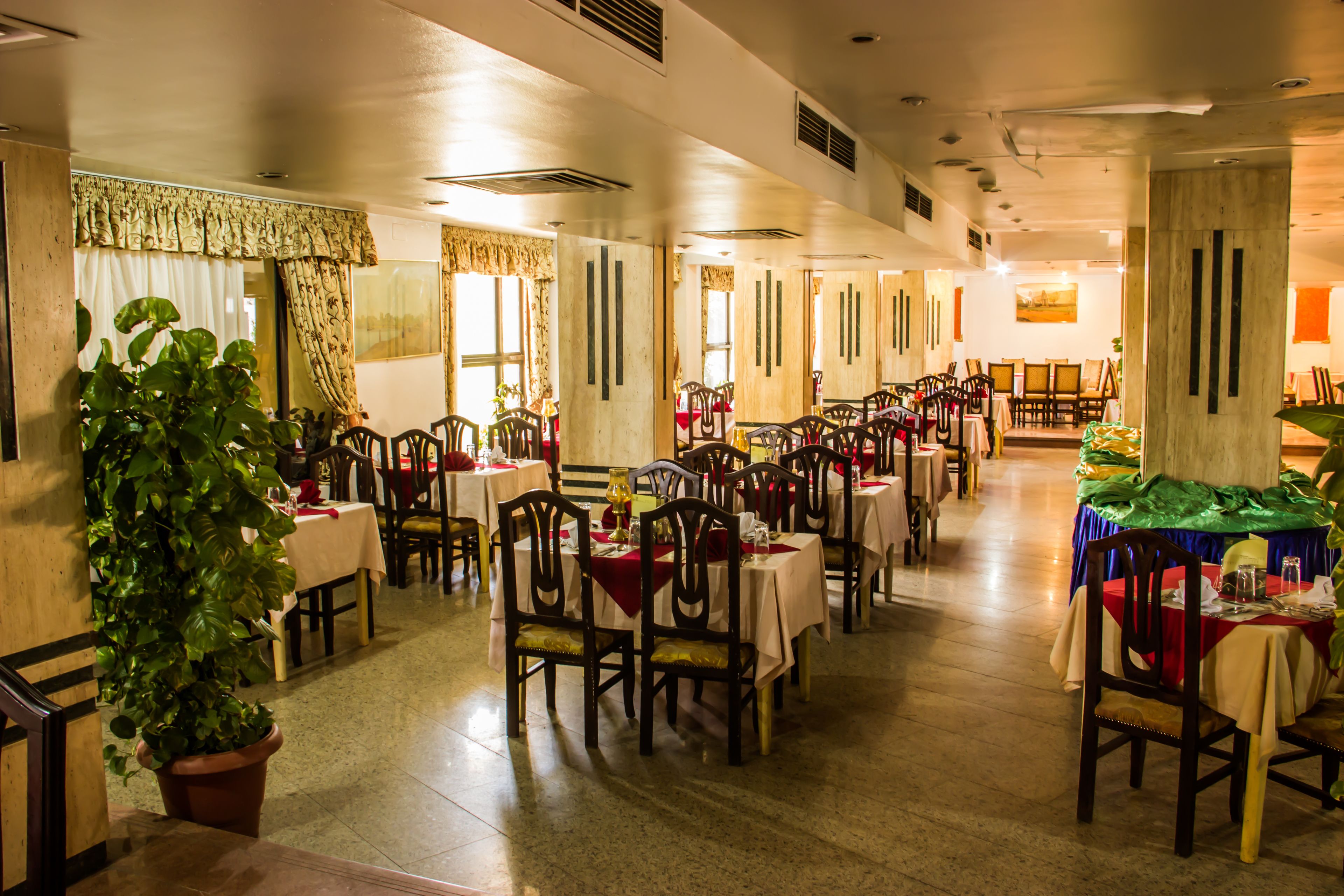 Un'ampia sala da pranzo con diversi tavoli e sedie, grandi finestre con tende e piante in vaso.