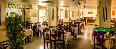 Un'ampia sala da pranzo con diversi tavoli e sedie, grandi finestre con tende e piante in vaso.