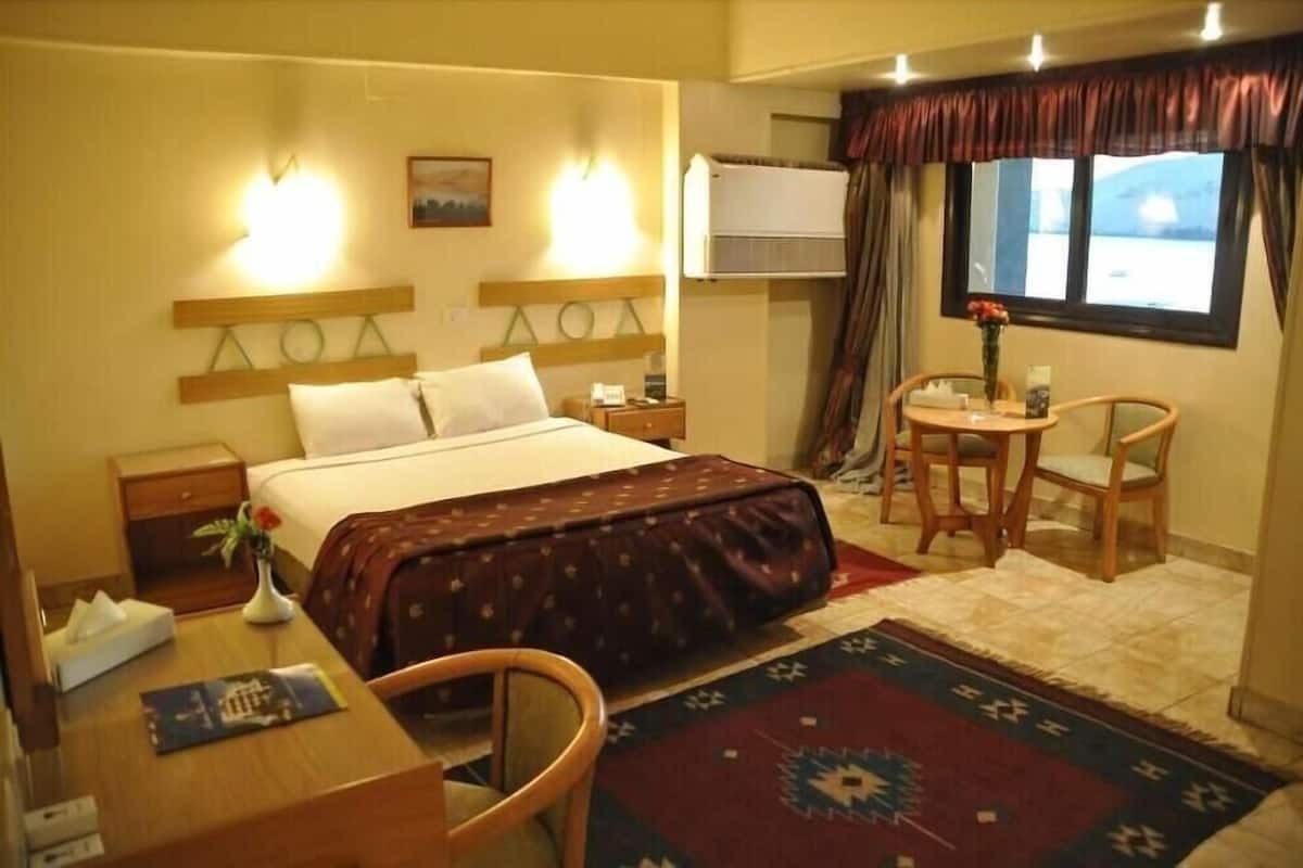 A hotel room with a bed, bedside tables, a desk, a chair, a small table with a vase, and a window with curtains.