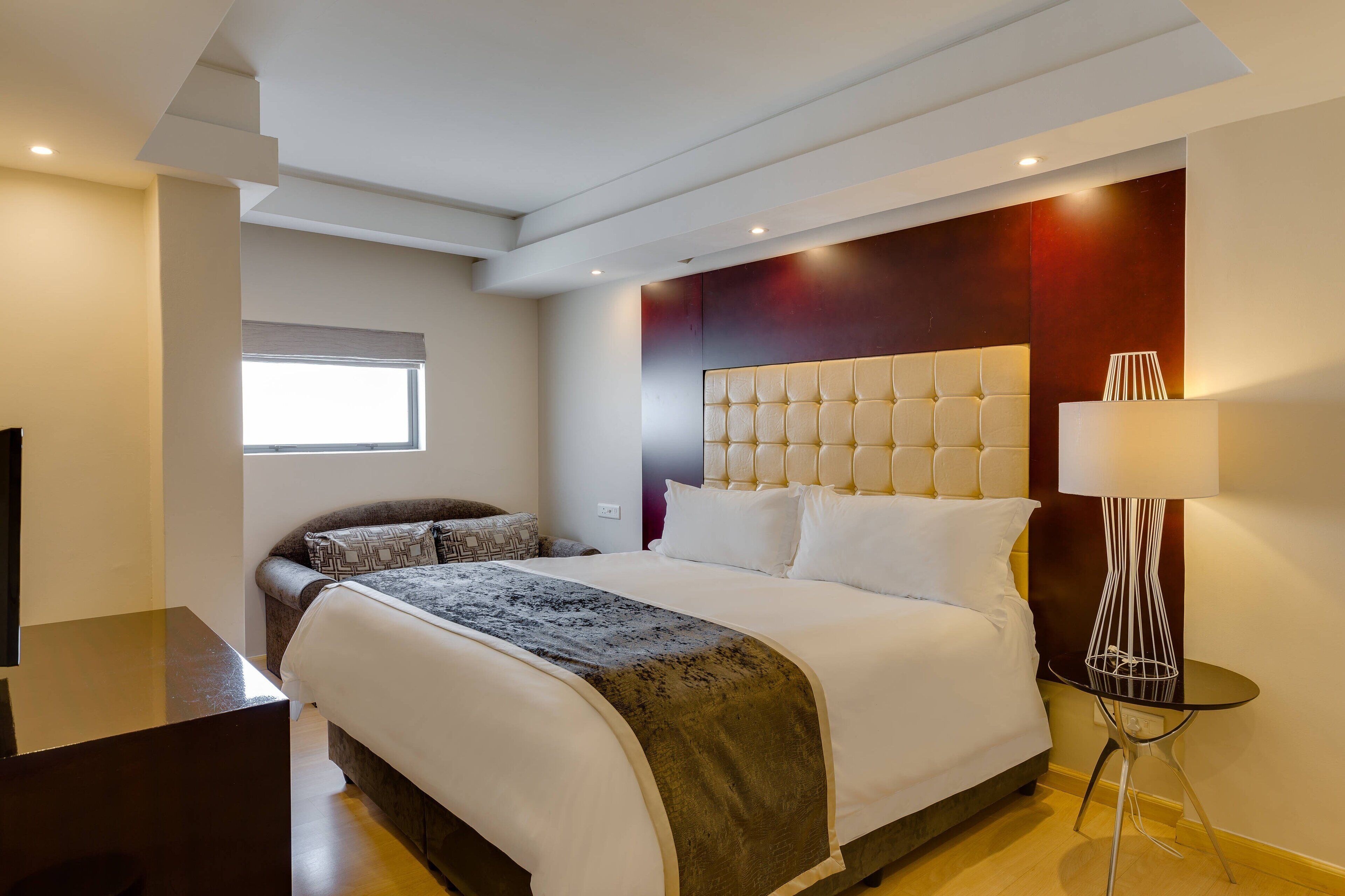 Una habitación de hotel moderna con una cama grande, una mesita de noche, una lámpara y un ventanal.