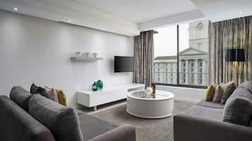 Uma sala de estar moderna com um sofá, uma mesa de centro redonda, uma televisão de tela plana e uma grande janela com vista para um edifício.