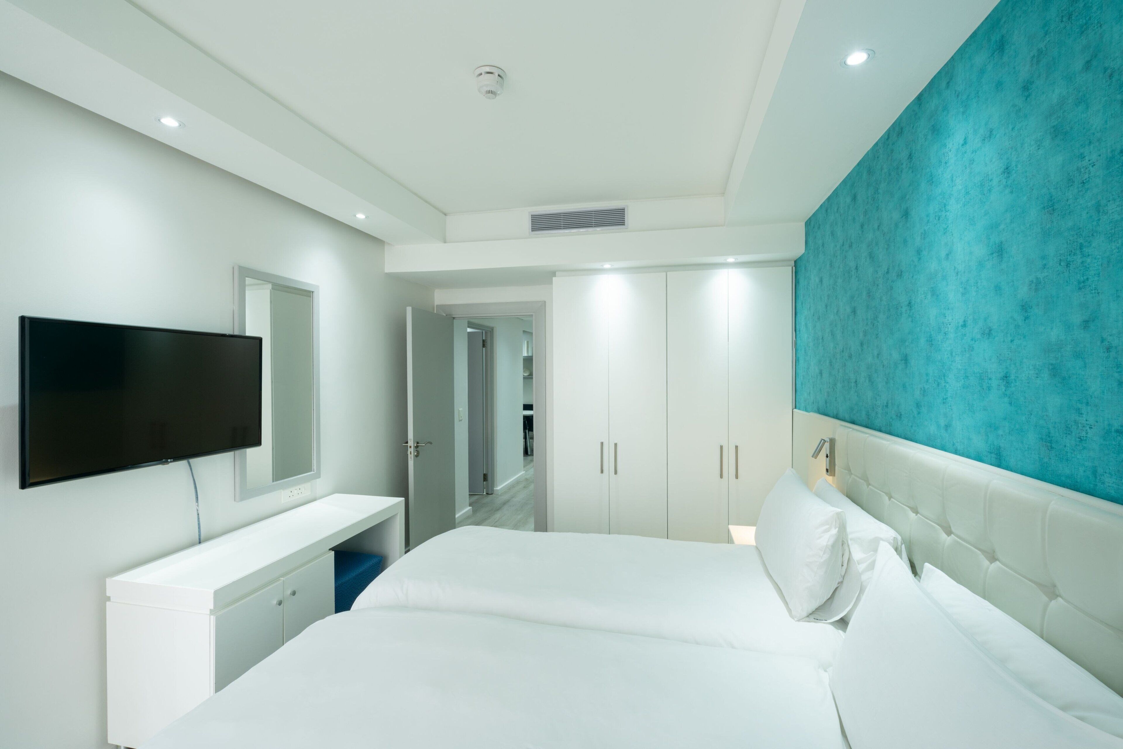 A modern hotel room with a large bed, a flat-screen TV mounted on the wall, a white bedside table, and a blue accent wall.