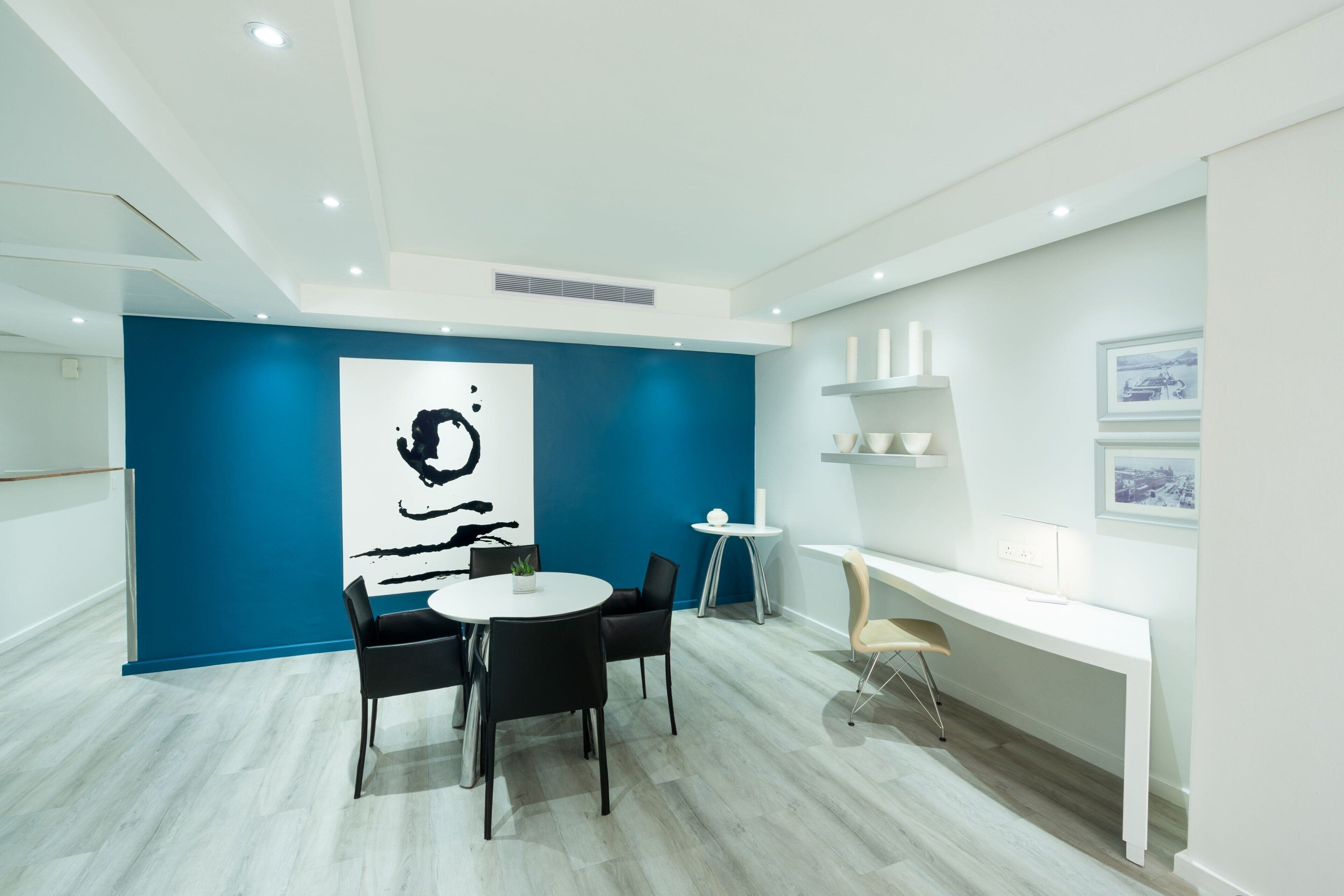 A modern office space with a blue accent wall, a round table with chairs, a white desk, and a large abstract painting.