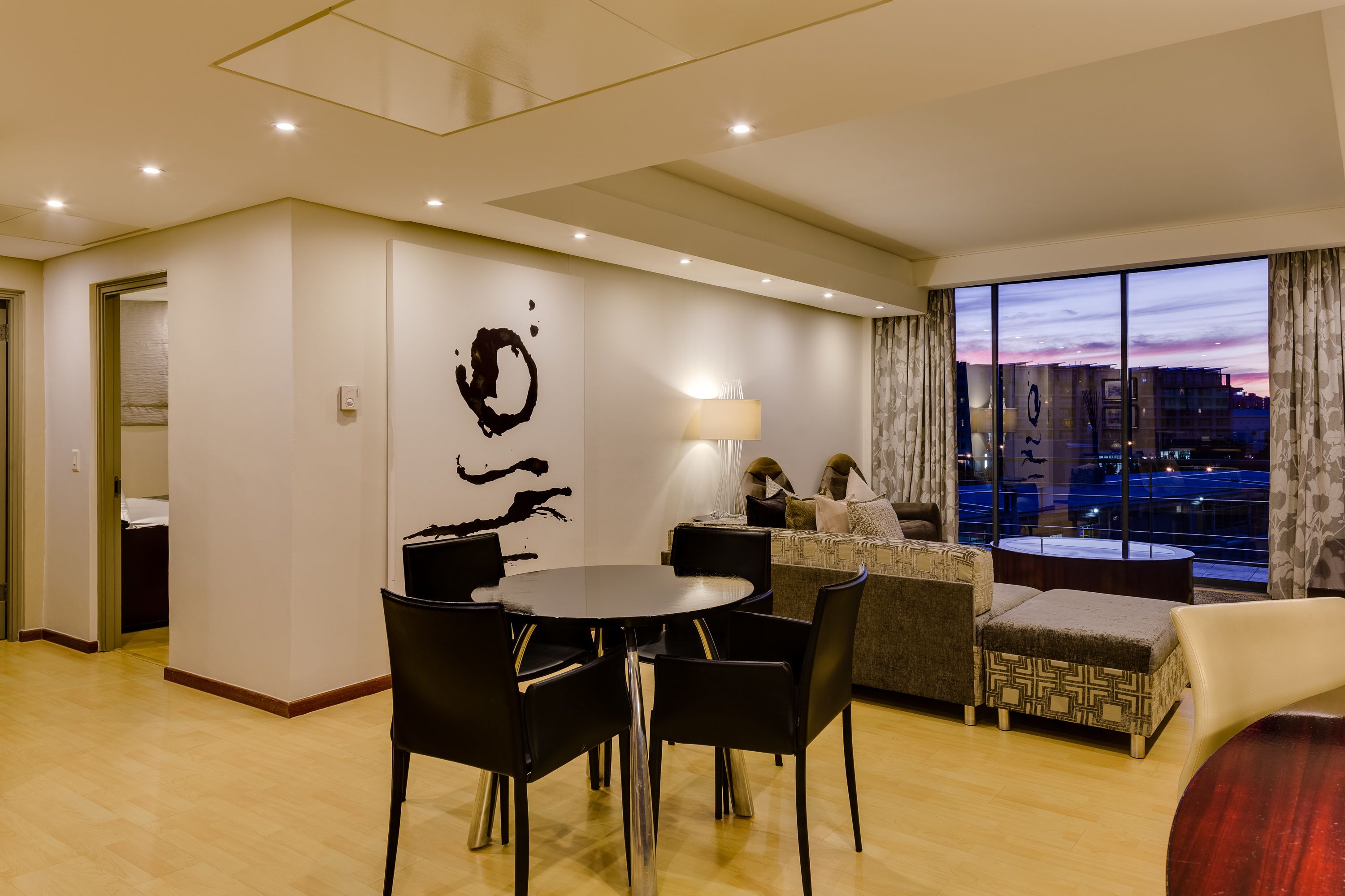 Et moderne hotelværelse med spiseplads, en sofa og et stort vindue med udsigt over byen.
