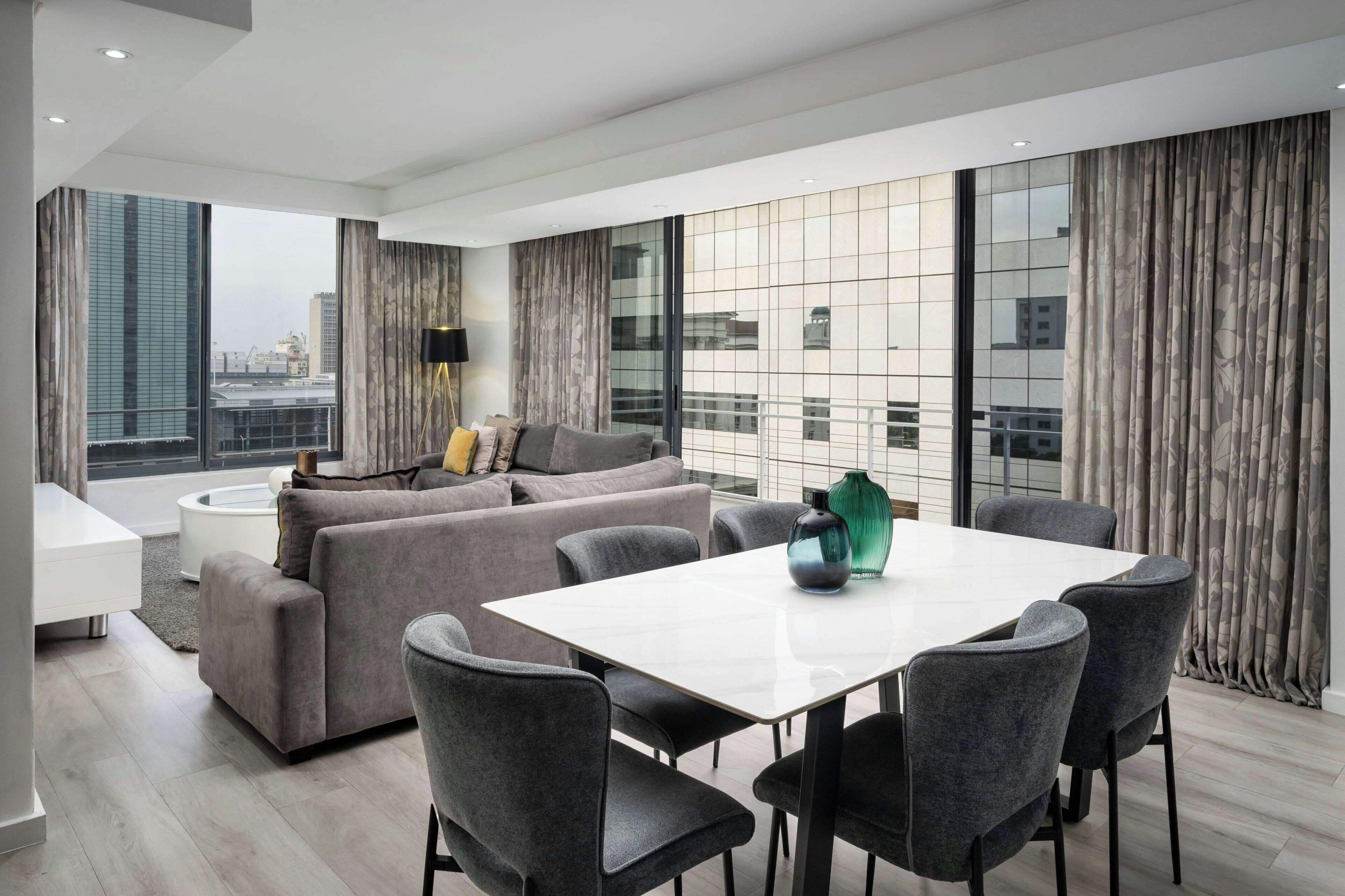 Een moderne woonkamer met een bank, eettafel en stoelen, grote ramen met gordijnen en uitzicht op de stad.