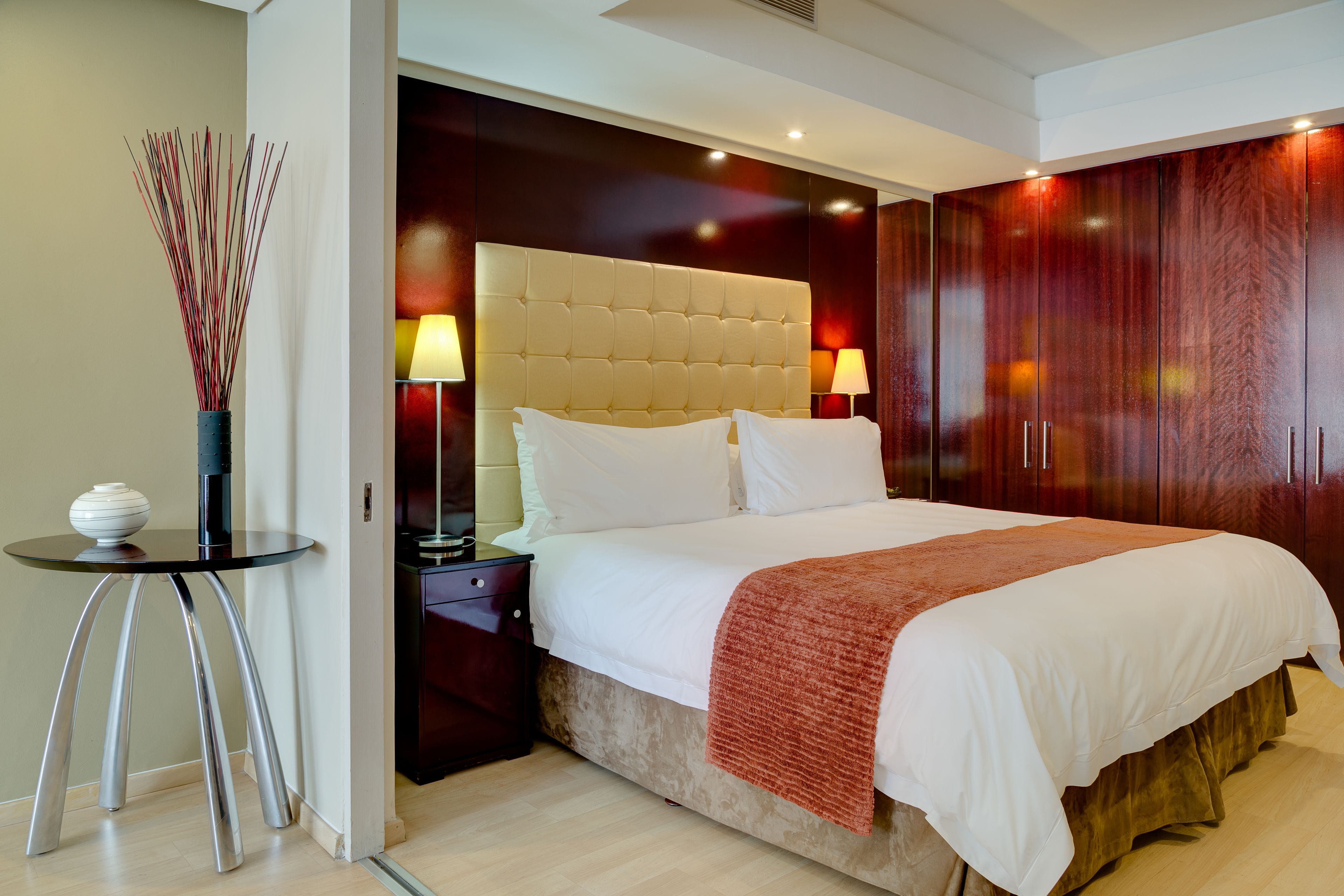 Ein modernes Hotelzimmer mit einem großen Bett, zwei Nachttischlampen, einem kleinen Tisch mit einer Vase und einem dunklen Holzschrank.