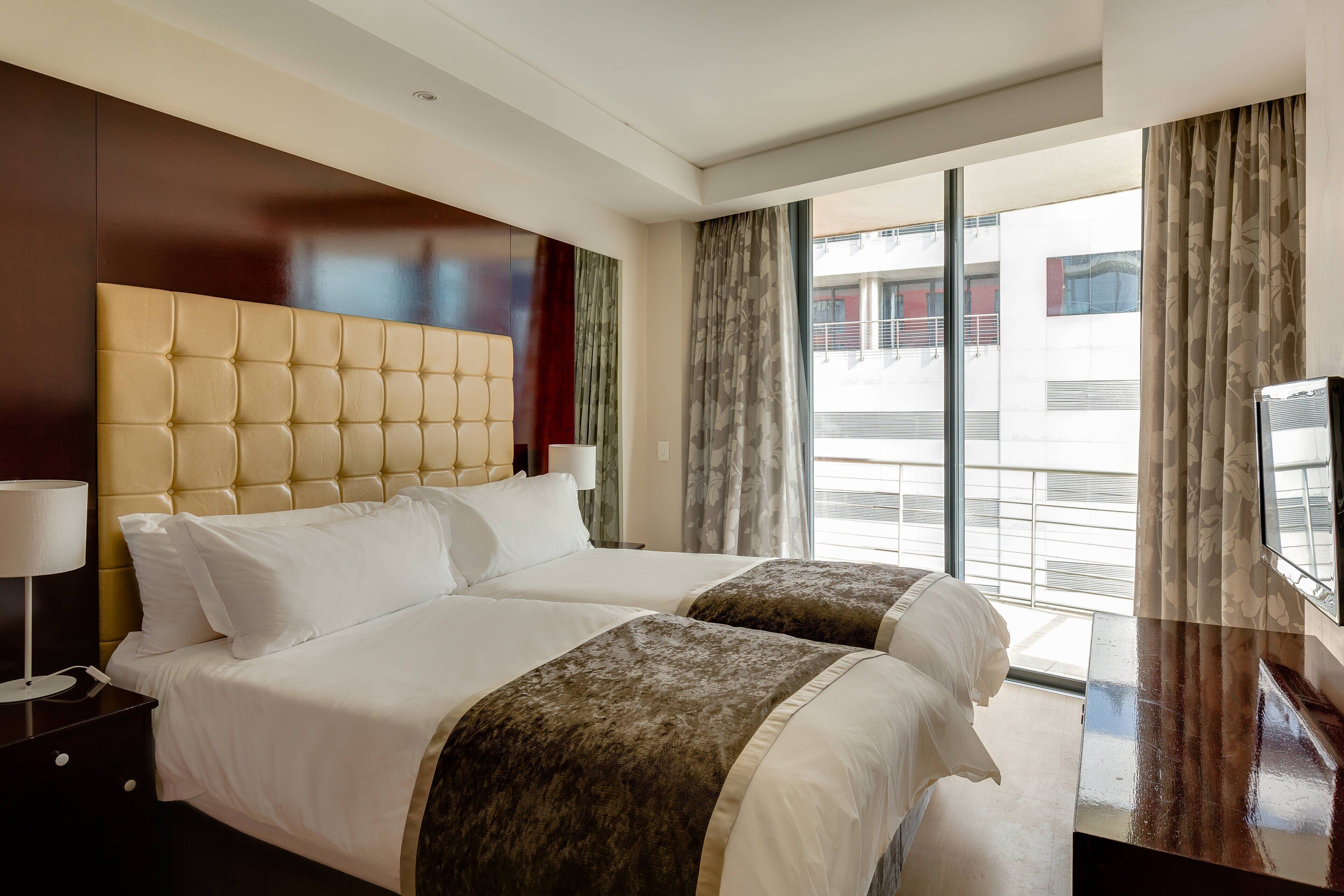 A hotel room with a large bed, two bedside tables, a TV, and a balcony with curtains.