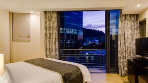 A hotel room with a bed, a TV, a desk, and a balcony with a city view.