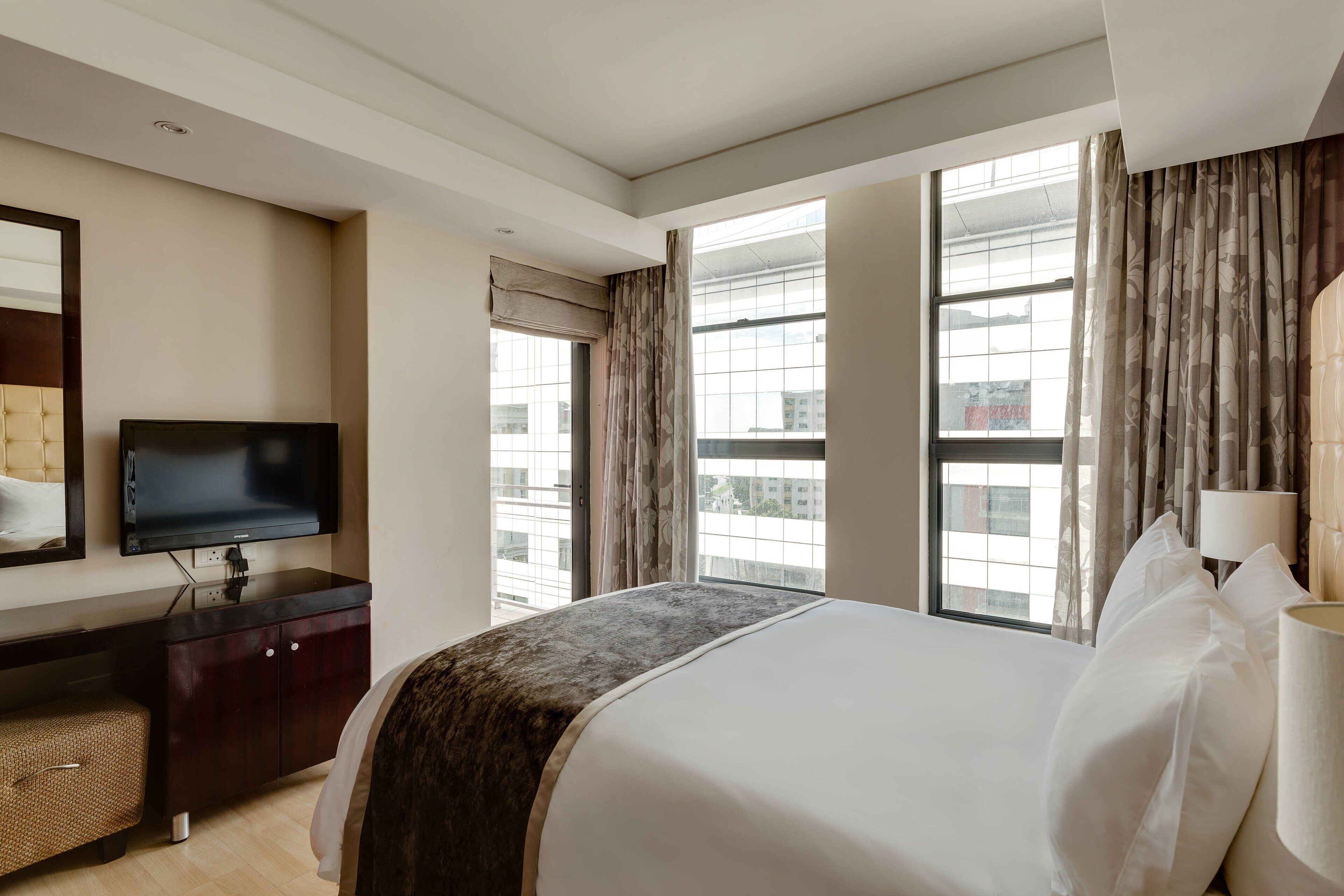 Une chambre d’hôtel avec un grand lit, un téléviseur posé sur une commode en bois foncé et de grandes fenêtres avec des rideaux.