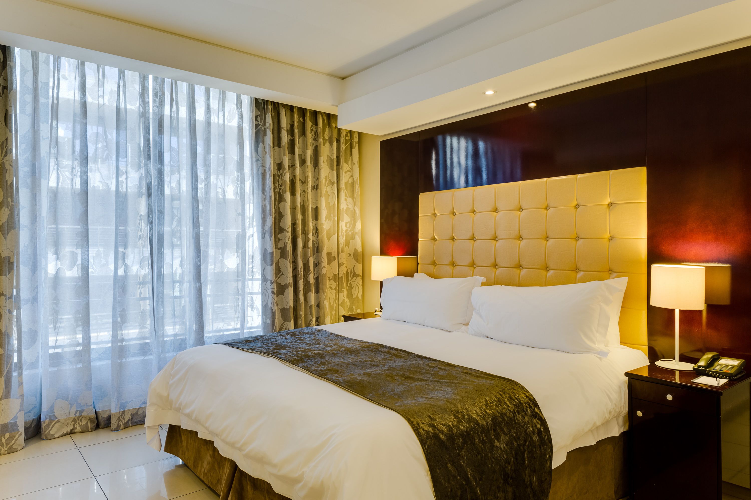 Ein Hotelzimmer mit einem großen Bett, zwei Nachttischlampen, einem Telefon und einem Fenster mit durchsichtigen Vorhängen.