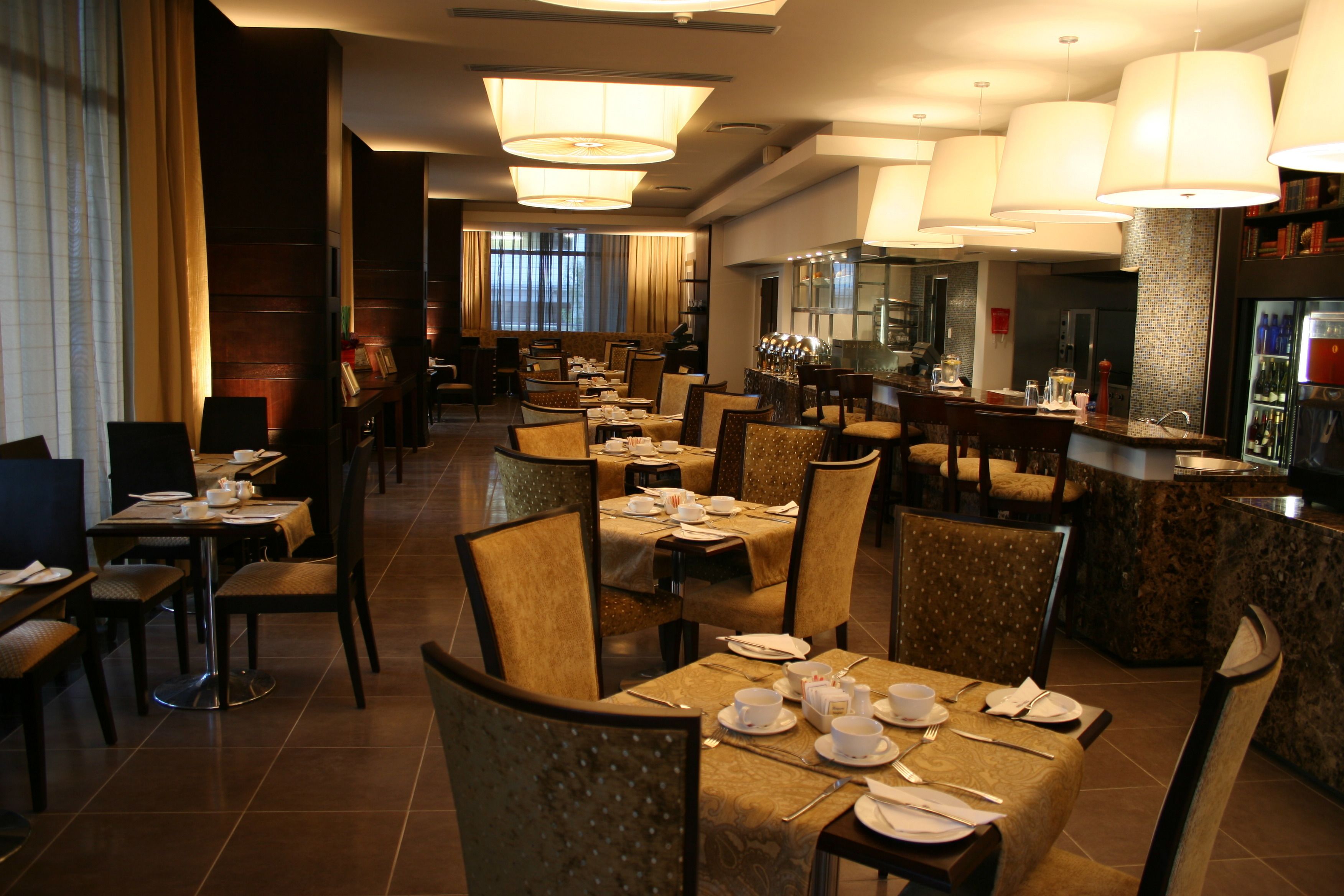 A restaurant with multiple dining tables and chairs, a bar area, and a view of the cityscape through large windows.