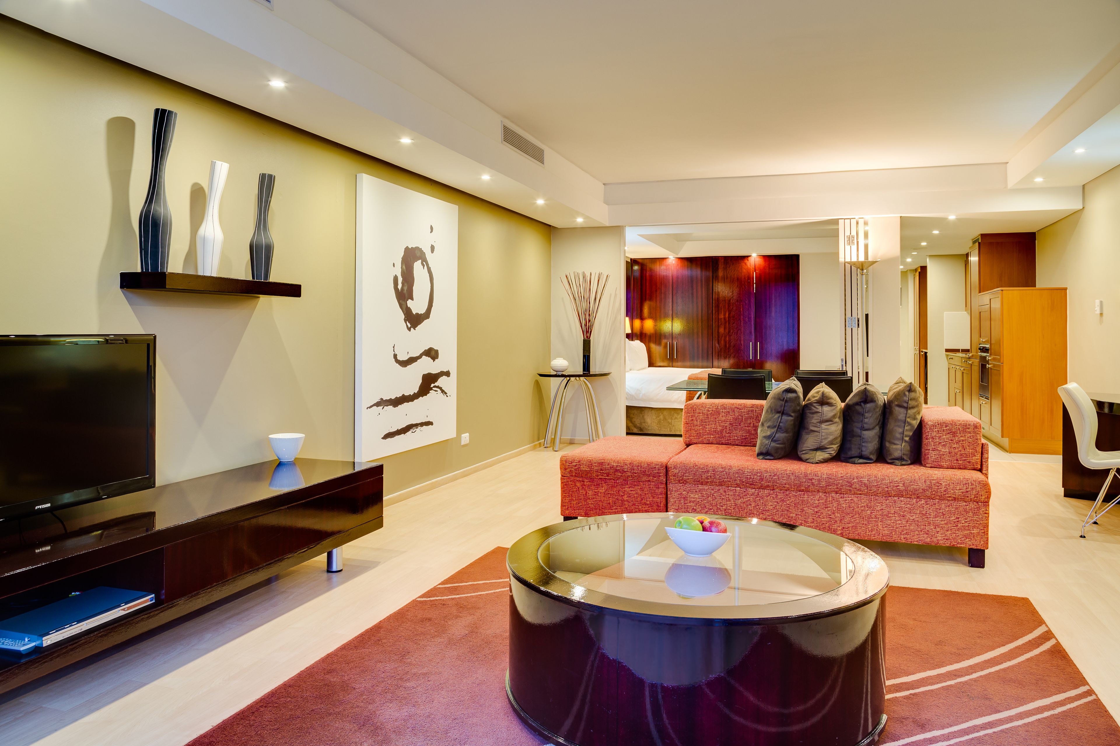 Una sala de estar moderna con un televisor de pantalla plana, un sofá rojo, una mesa de centro de vidrio y un comedor al fondo.