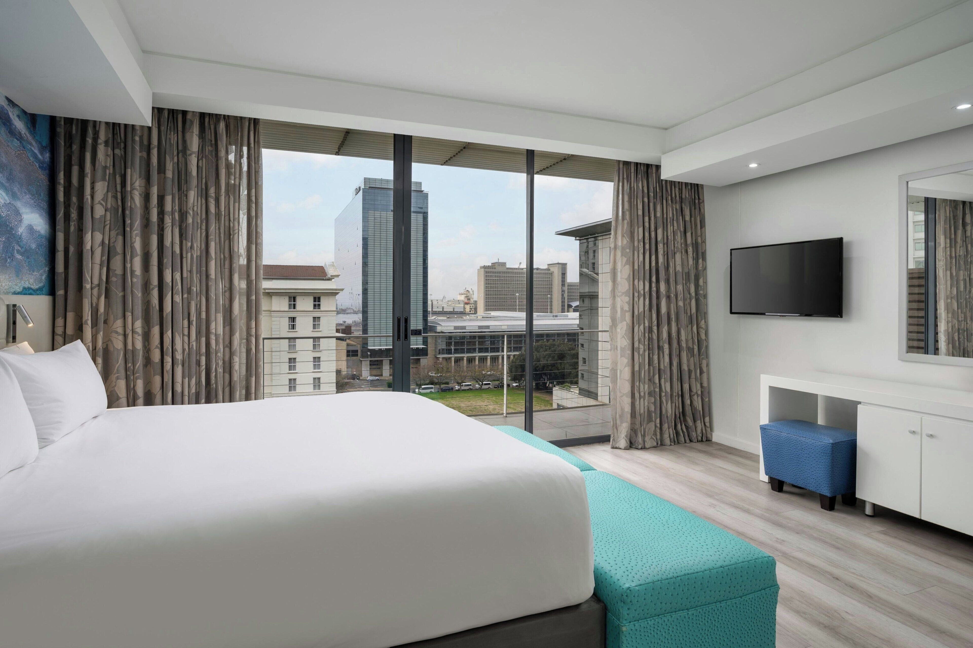 Ein Hotelzimmer mit einem großen Bett, einem Fernseher, einem Schreibtisch und Blick auf Stadtgebäude.
