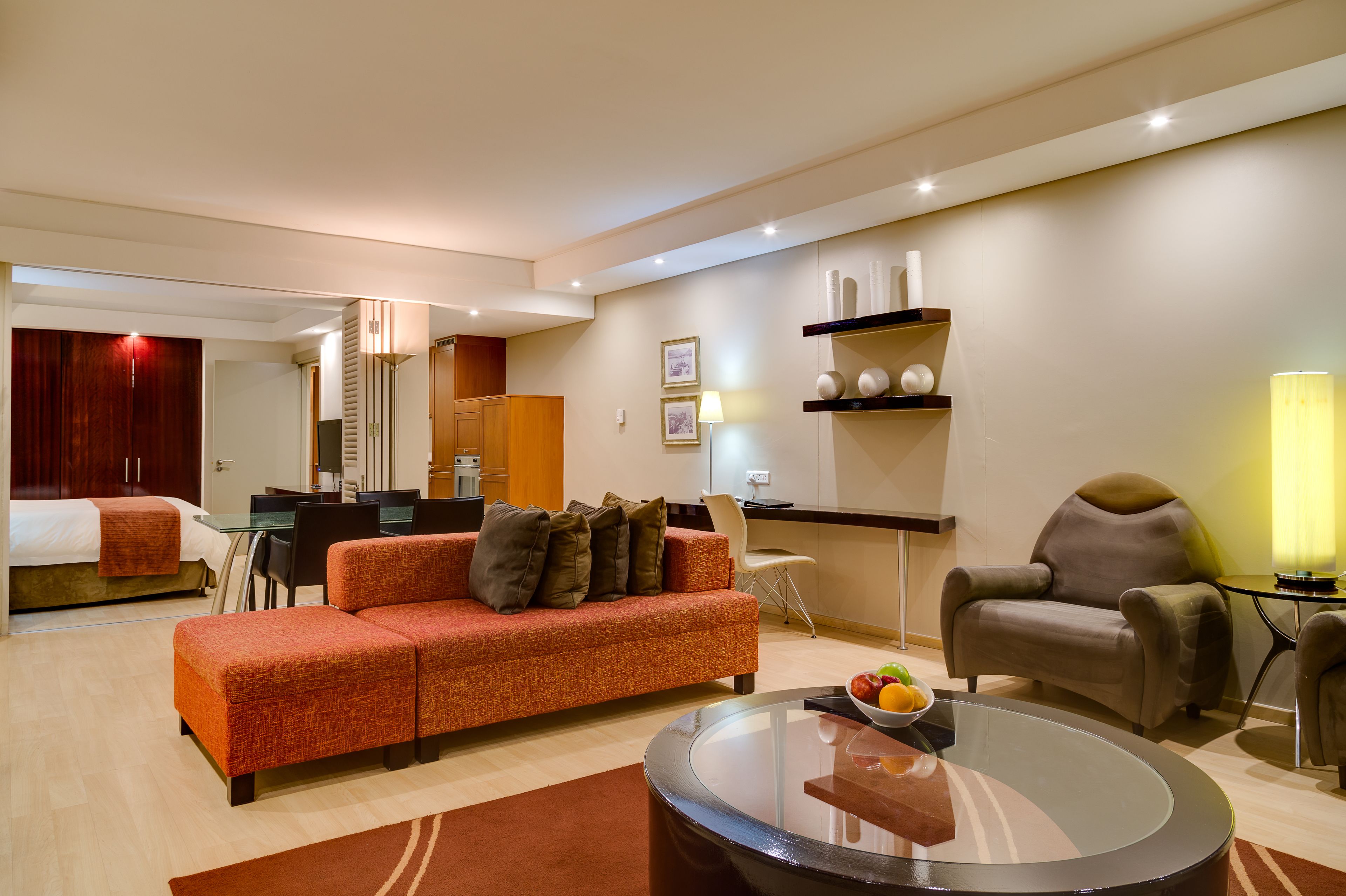 A modern hotel room with a sofa, armchair, dining table, and a glass coffee table.