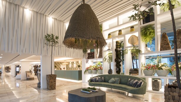 Een ruime lobby met een modern ontwerp, met een grote geweven lamp, een gebogen zithoek en diverse planten en decoratieve items op planken.