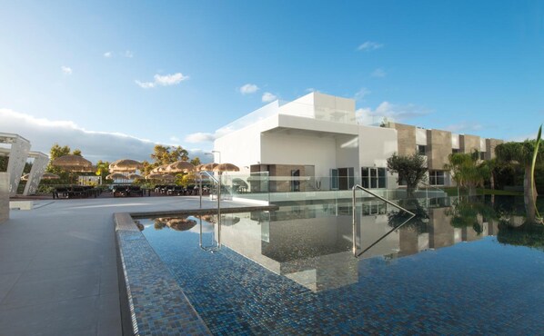 Un hotel moderno con una gran piscina exterior, una zona de estar cubierta y una vista despejada del paisaje circundante.