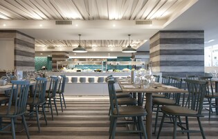 Un intérieur de restaurant moderne avec un plancher en bois, des chaises bleues et un comptoir central où est présenté un buffet.