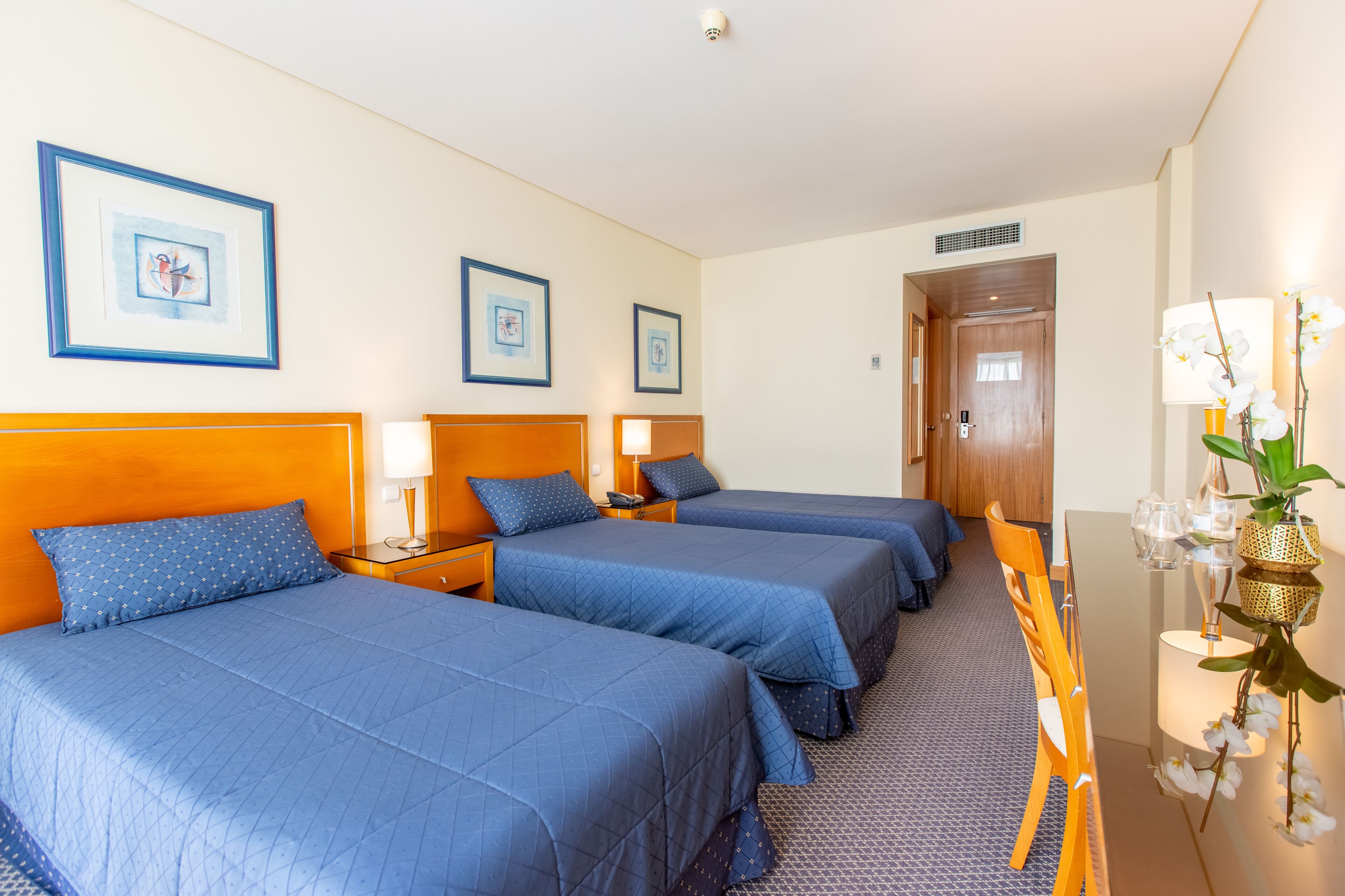 A hotel room with three beds, a wooden headboard, a desk with a chair, and framed artwork on the wall.