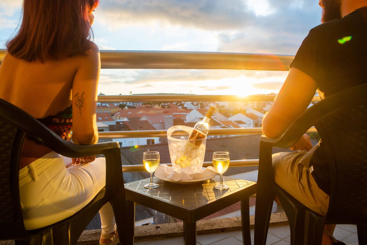 Deux personnes sont assises à une table, une bouteille de champagne et deux verres devant elles, sur un balcon donnant sur une vue panoramique de la ville.