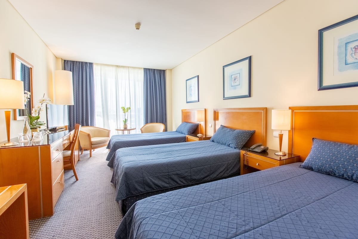 Une chambre d’hôtel avec trois lits, un bureau, une chaise et une petite table avec une lampe.