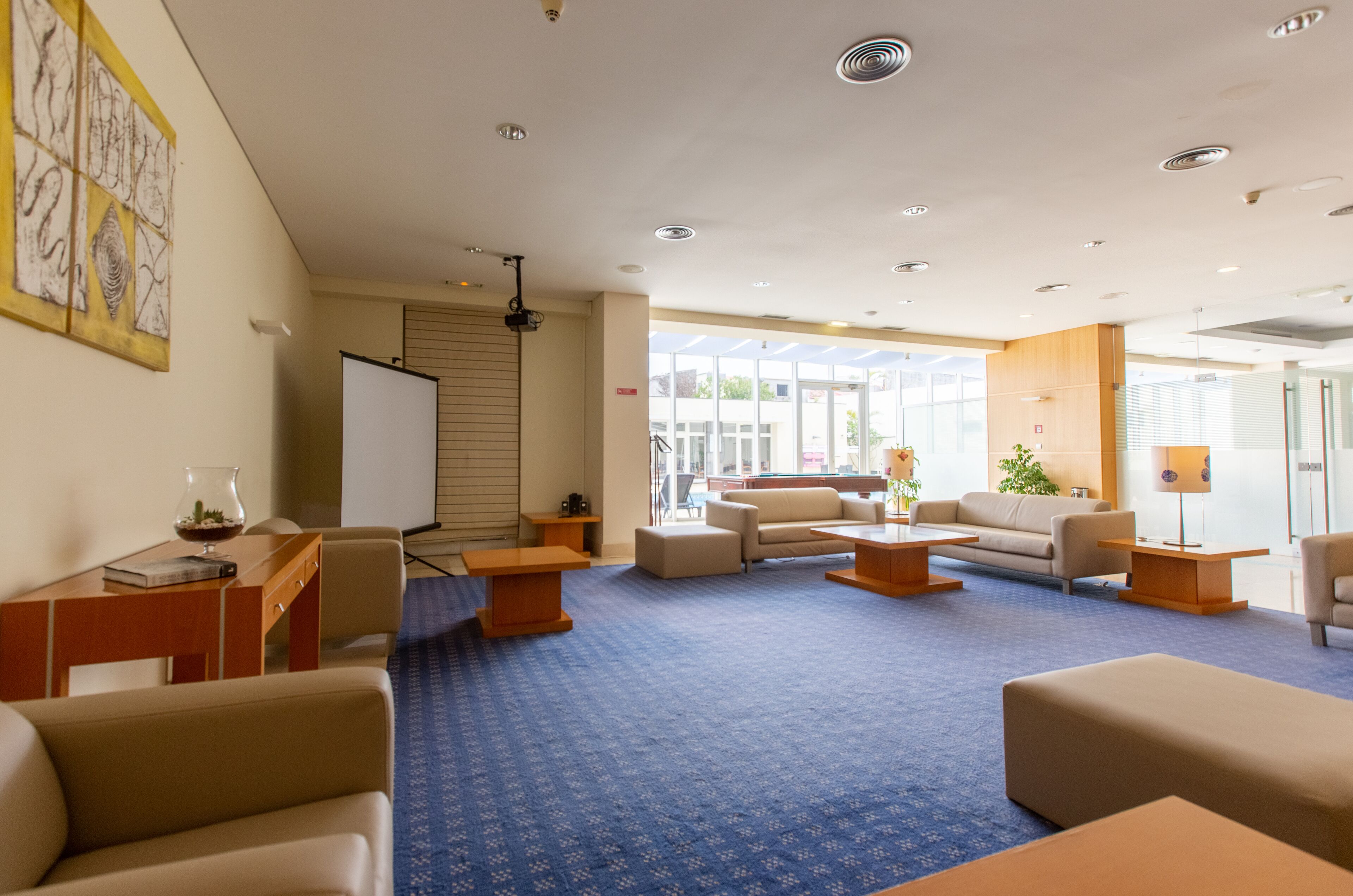 A spacious lobby with a blue carpet, multiple seating areas, a whiteboard, and a large window providing natural light.