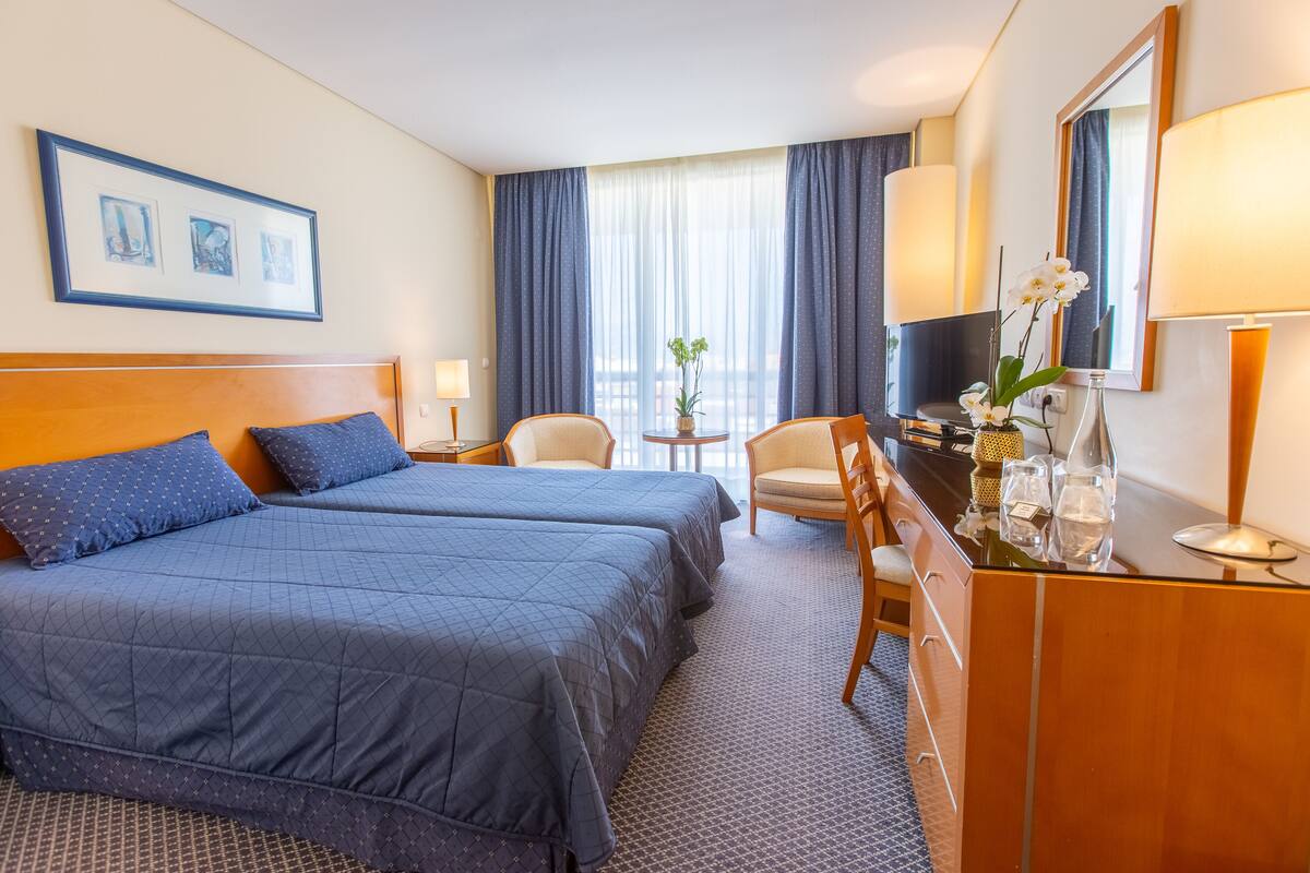 Une chambre d’hôtel avec deux lits, un bureau, une chaise, une petite table ornée d’un vase de fleurs et un grand miroir.