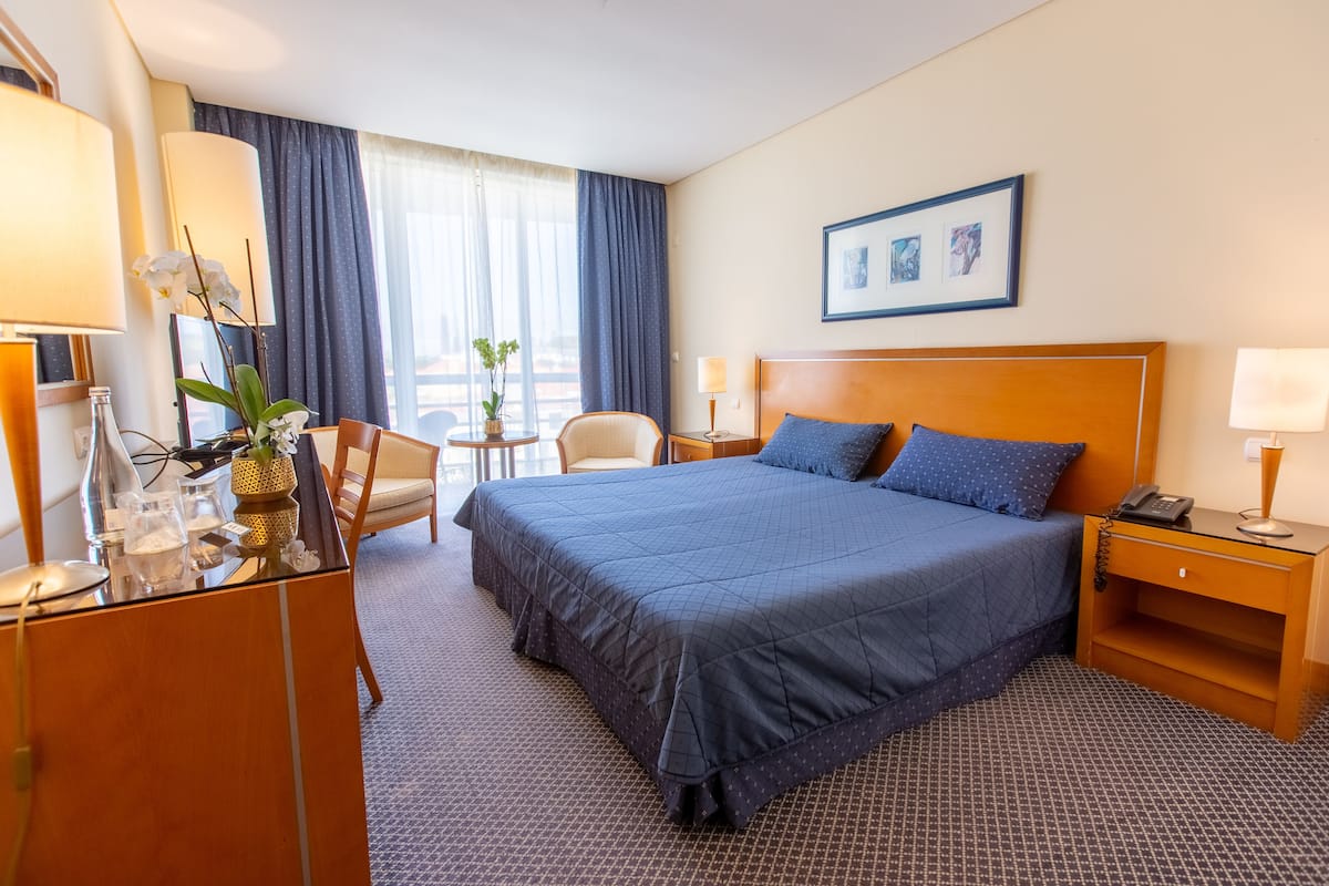 Une chambre d’hôtel avec un grand lit, un bureau avec une lampe, une chaise, une petite table avec une plante et une fenêtre avec des rideaux.