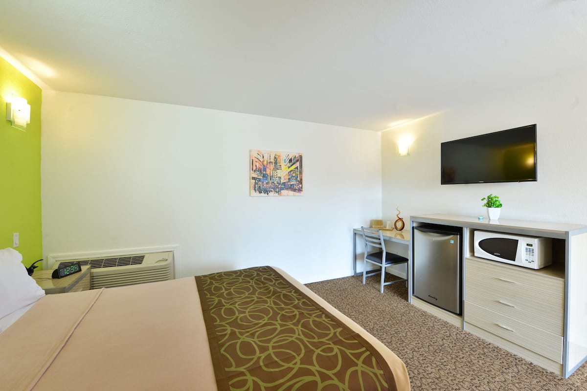 A hotel room with a bed, a desk with a chair, a microwave, a refrigerator, and a flat-screen TV mounted on the wall.