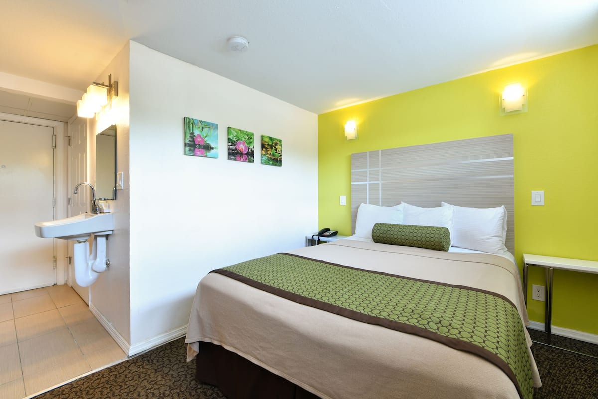 A hotel room with a bed, a bedside table, a nightstand, a mirror, a sink, and a door.