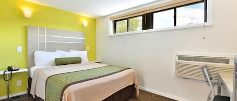 A hotel room with a bed, a chair, a desk, a window, and a wall-mounted heater.