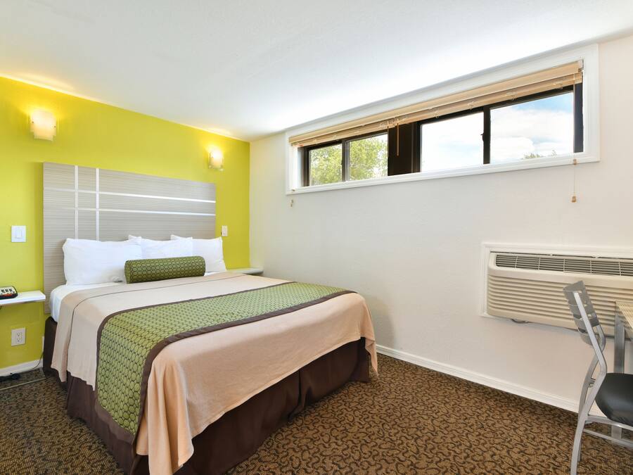 A hotel room with a bed, a chair, a desk, a window, and a wall-mounted heater.