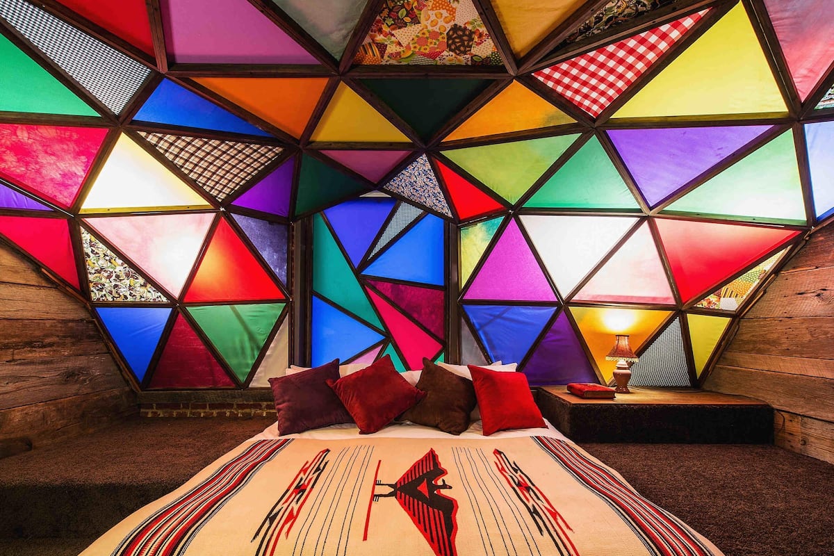 A room with a geometric-patterned ceiling, a bed with a patterned bedspread, and a nightstand with a lamp.