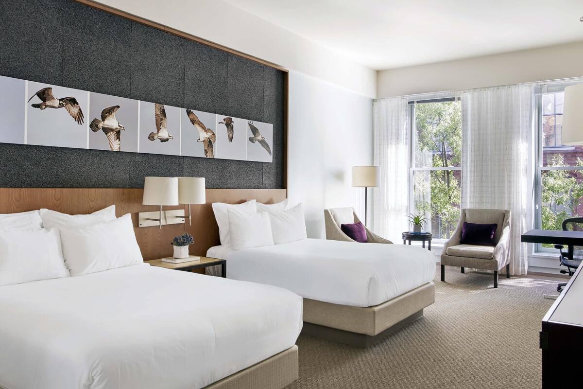 A hotel room with two beds, a desk, a chair, a lamp, and a large window with a view.