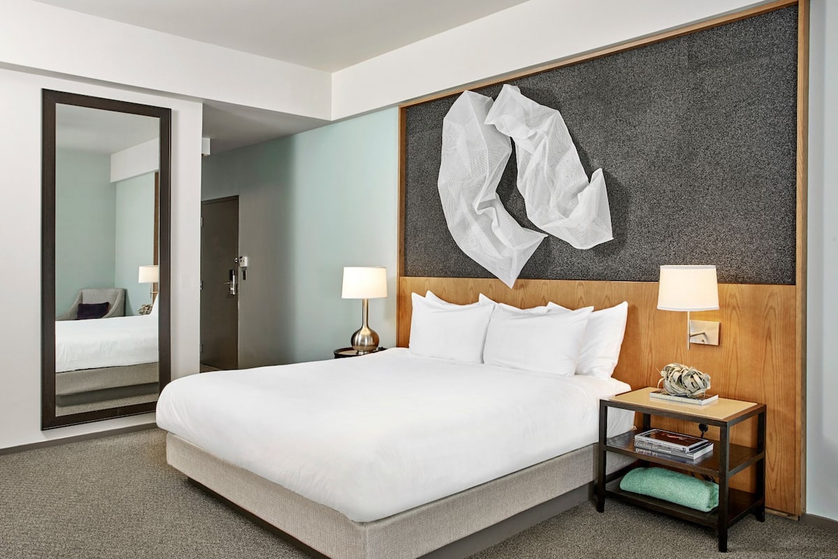 A hotel room with a large bed, two bedside tables with lamps, a wooden headboard, a large mirror, and a wall-mounted abstract art piece.