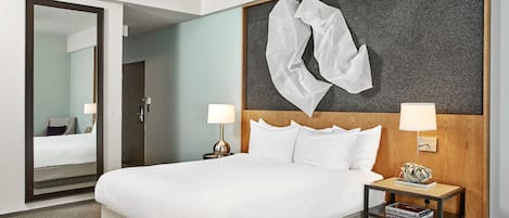 Habitación de hotel con una cama grande, dos mesitas de noche con lámparas, un cabecero de madera, un espejo grande y una obra de arte abstracto colgada en la pared.