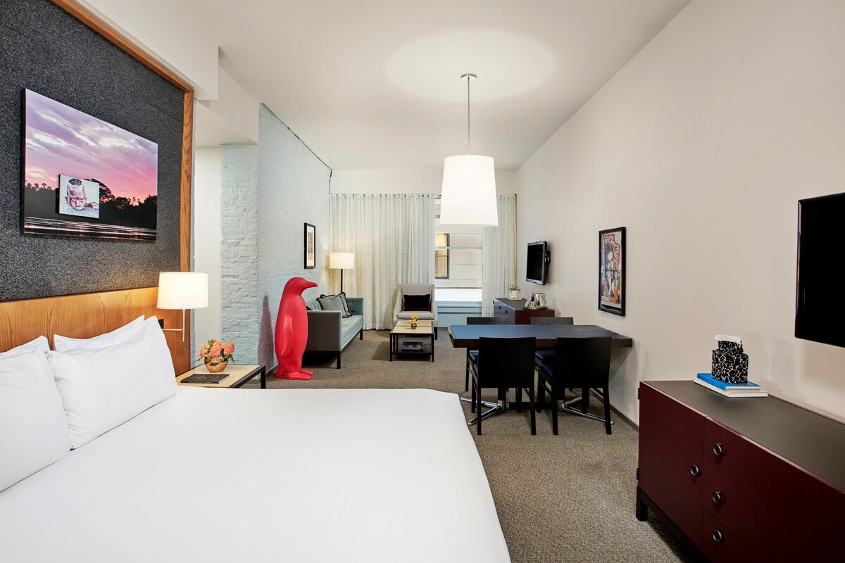 A hotel room with a large bed, a desk, a chair, a red sculpture, a TV, and a painting on the wall.