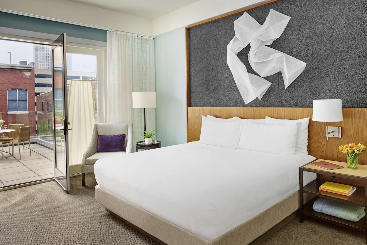 A hotel room with a large bed, a nightstand with a lamp, a chair, and a view of the outdoors.