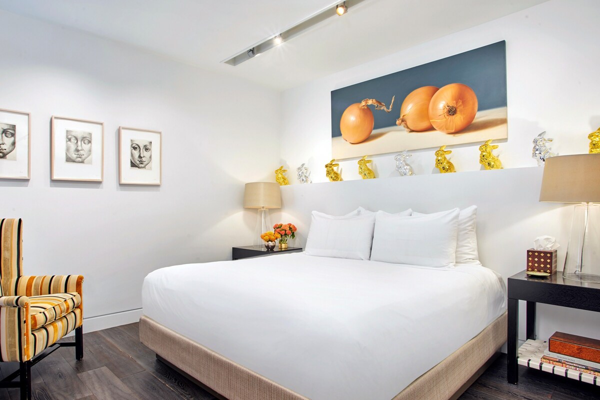 A hotel room with a large bed, a striped armchair, a nightstand with a lamp, and wall art including framed pictures and a painting of onions.