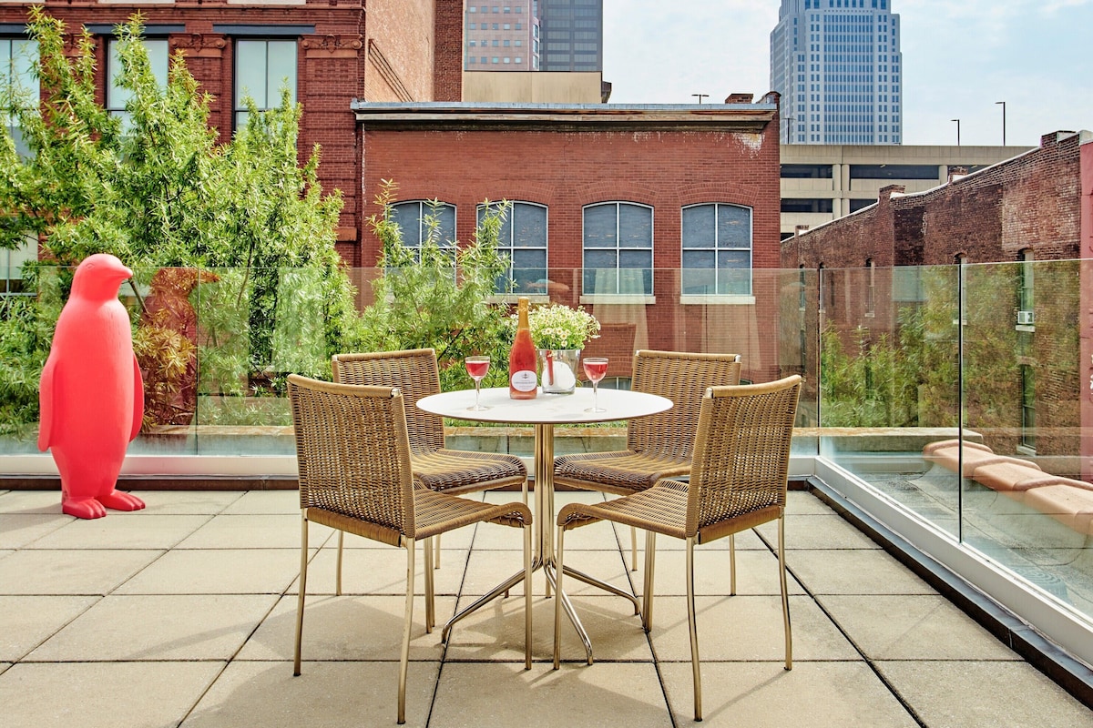 A rooftop terrace with a round table, wicker chairs, a red sculpture, and a view of city buildings.