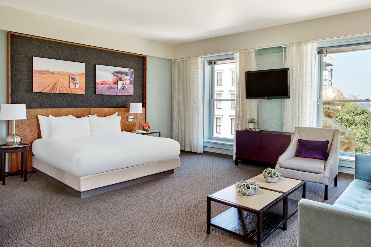 A hotel room with a large bed, a TV mounted on the wall, a desk with a lamp, and a view of the city through large windows.