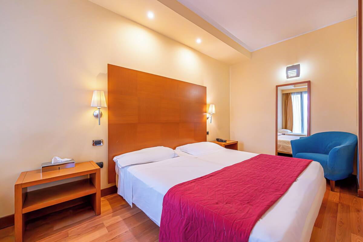 A hotel room with a large bed, bedside lamps, a wooden nightstand, a blue armchair, and a mirror on the wall.