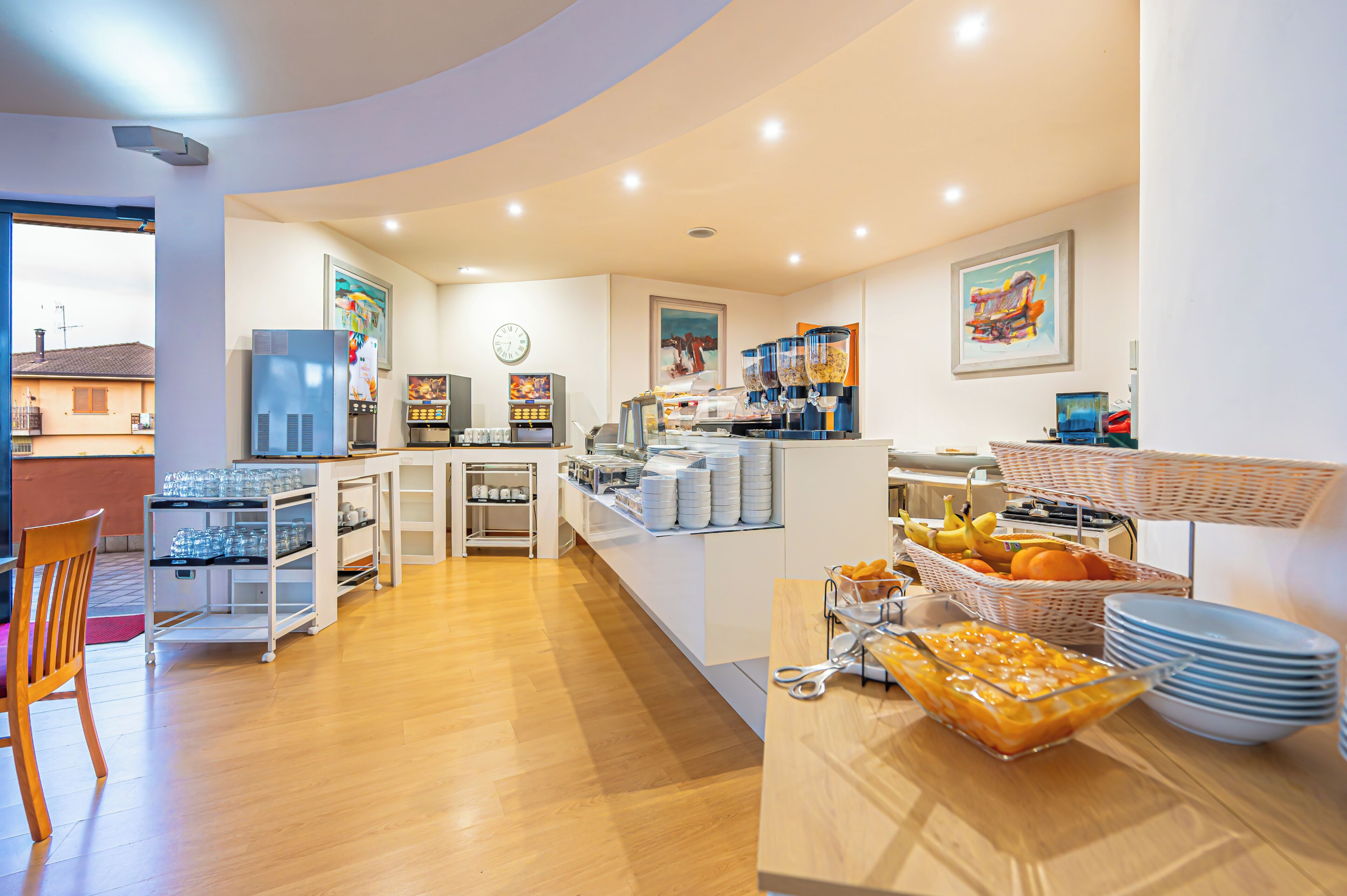 A breakfast area with a refrigerator, coffee machines, and a buffet with fruits and dishes.