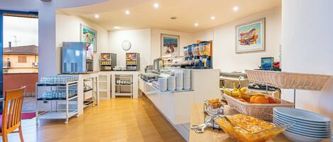 A breakfast area with a refrigerator, coffee machines, and a buffet with fruits and dishes.