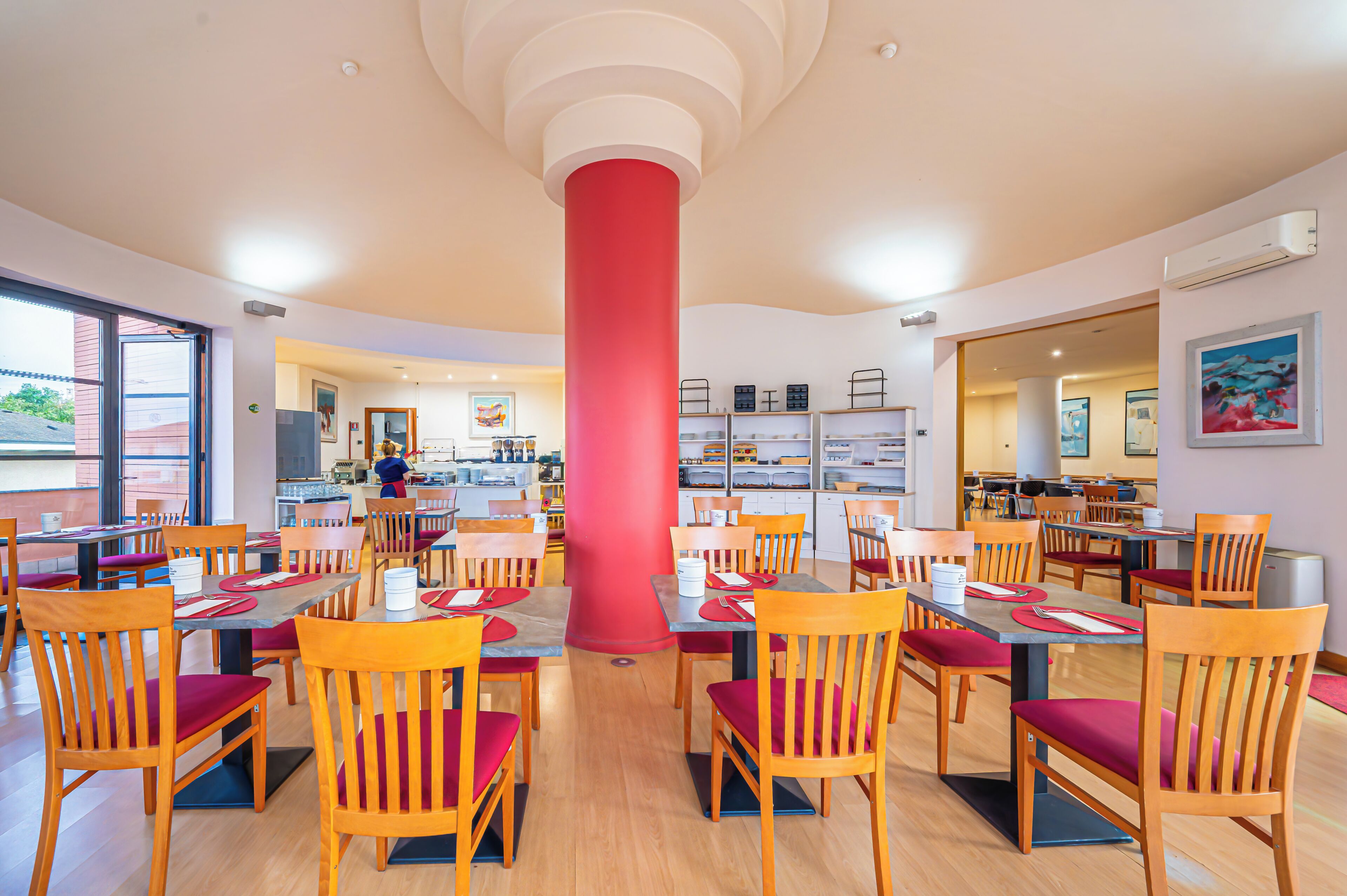 A spacious restaurant with wooden tables and chairs, a central red pillar, and a view of the kitchen area.